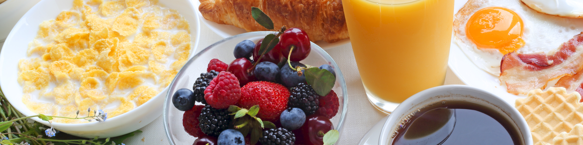 A hearty breakfast spread with waffles, croissants, eggs, yogurt, fruit bowl, juice, and coffee.