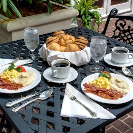 Outdoor breakfast on a patio table with bacon, eggs, croissants, champagne flutes, and coffee cups.