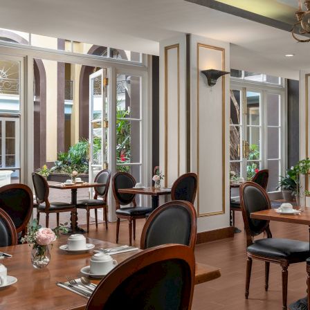 An elegant caf&eacute; dining room with wooden chairs, round tables set for tea, and tall arched windows beyond ornate interior mirrors.