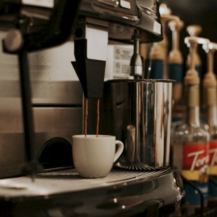 A coffee machine pours hot coffee into a white cup on the counter, with other drinks and bottles blurred in the background.