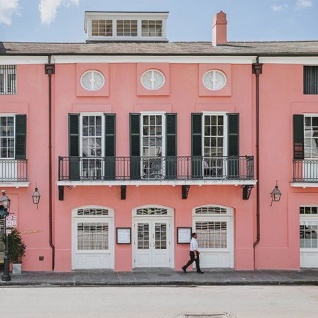 A bright pink building fa&ccedil;ade with balconies, shuttered windows, and a man walking by on the street.
