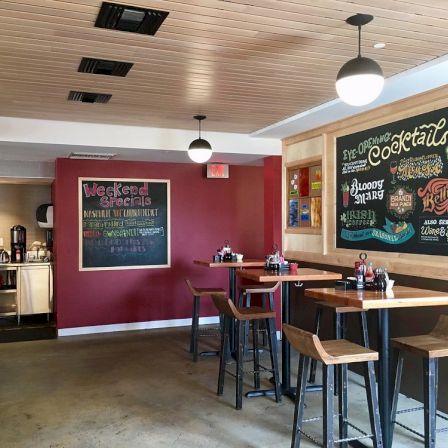 A cozy bar interior with stools and hanging lights, featuring large chalkboard menus for cocktails and weekend specials.