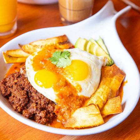 A white plate of fried eggs over savory meat and roasted potatoes, garnished with cilantro, with orange drinks in the background.
