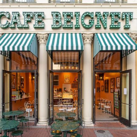 Cafe Beignet storefront with green-and-white striped awnings and outdoor tables in front of the columns.