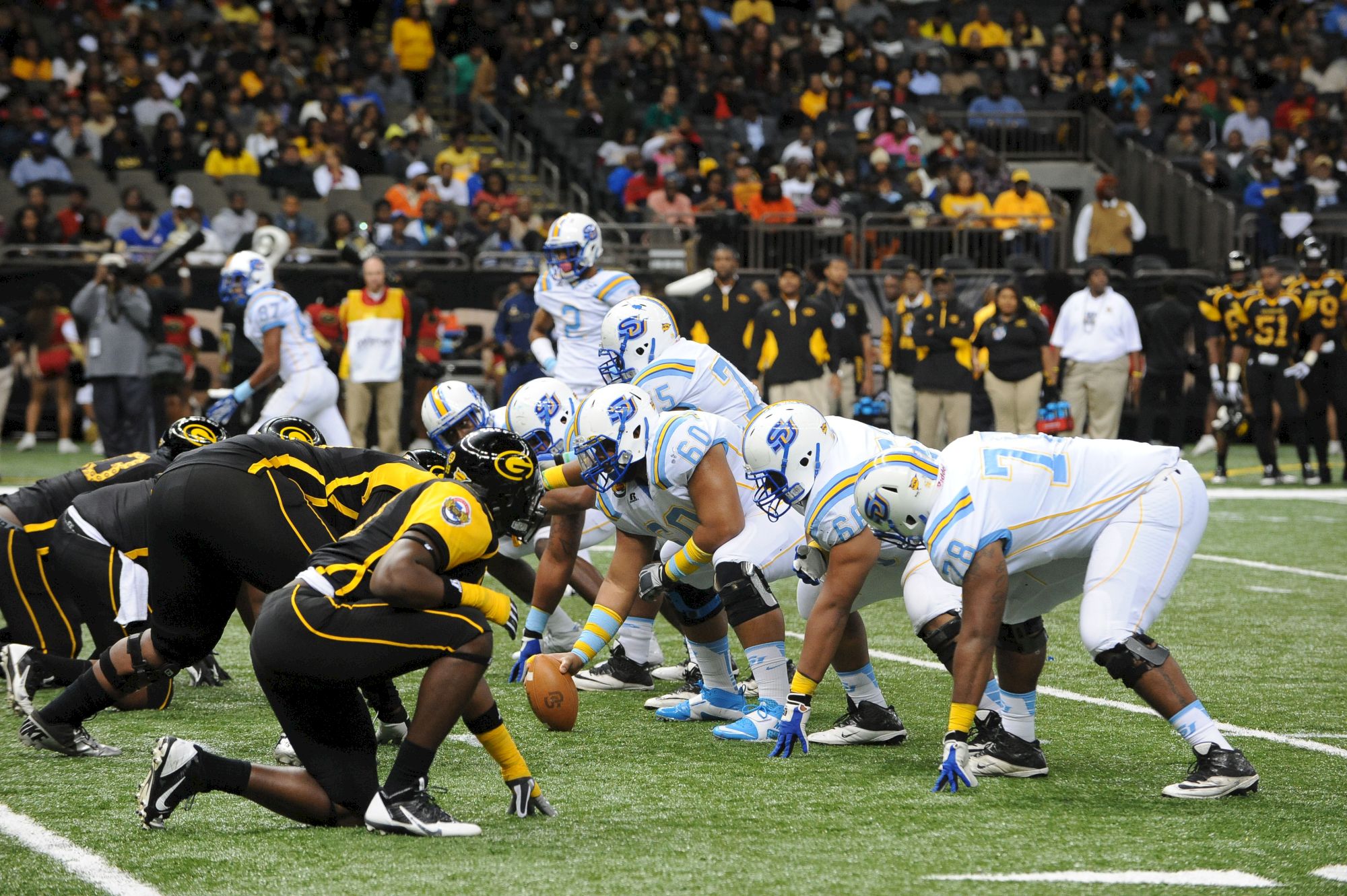 Football players in white/light-blue and black/yellow crowd a scrum near the line, with coaches and fans in the stands.