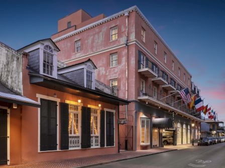 A row of historic buildings with balconies and flags along a street at dusk, including a pink facade and lit windows.