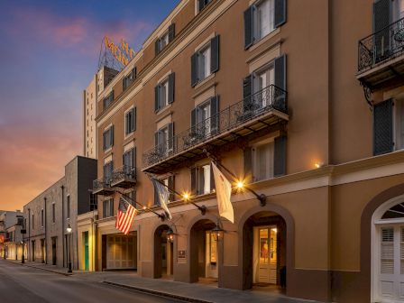 Historic hotel facade with balconies and warm lights at dusk, plus American flags and a neon sign on the street.