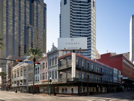 Downtown skyline with palm-lined street, a vertical &ldquo;RUBENSTEINS&rdquo; sign, and a billboard reading &ldquo;CANALI&rdquo; above storefronts and hotels.
